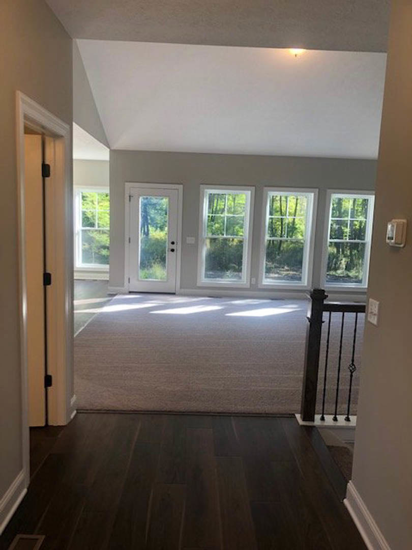 Hallway with dark wood flooring and carpet, black metal staircase railing, white door with window, large windows showing trees outside, white wall fixture.
