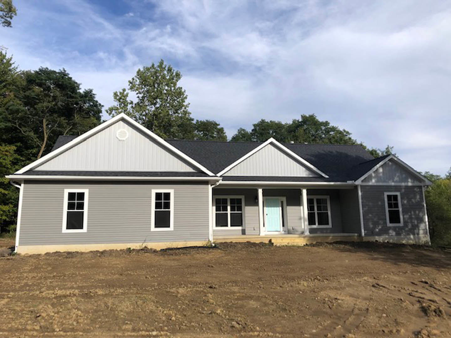 Partially built home with exposed framing, blue front door, white framed window, white door with black handle, dirt yard with white pole, surrounding trees, and small white cottage