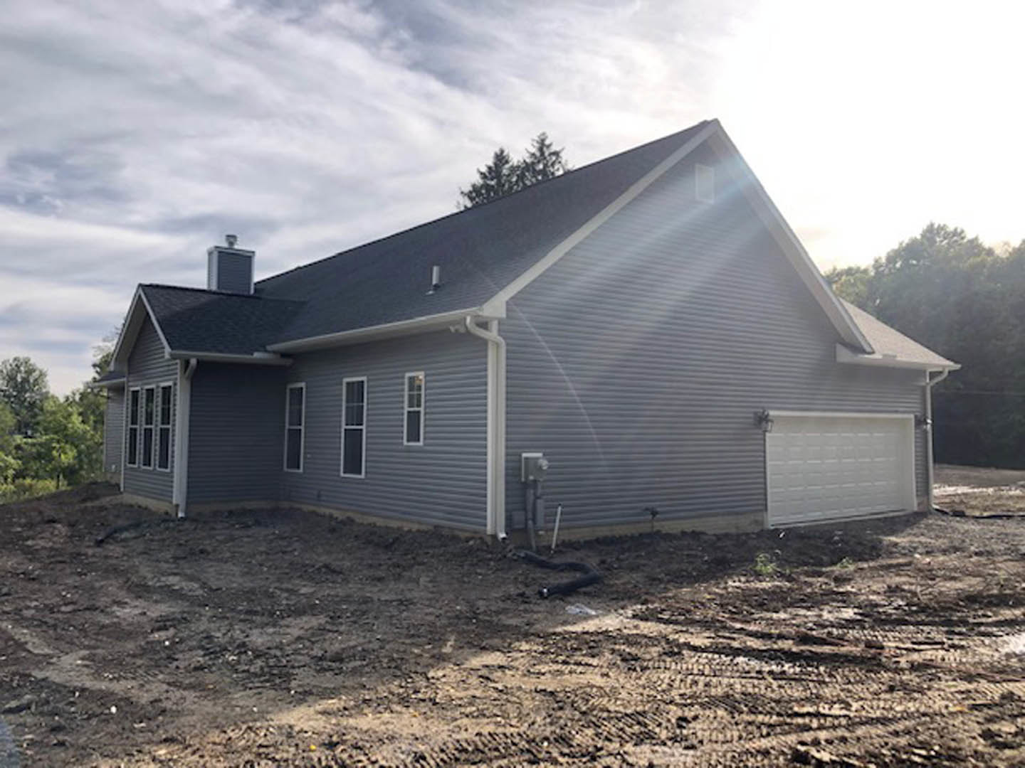 Two-story house under construction with exposed framing, dirt yard, white garage door, sprinkler system pipes, and large windows; man standing near garage.