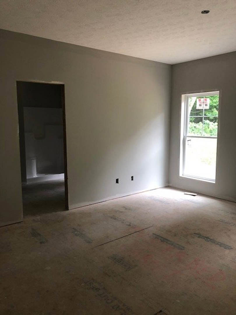 White plaster walls, open wooden door, large window with a view of green trees, light-colored flooring with black and red text, ceiling with recessed lighting.