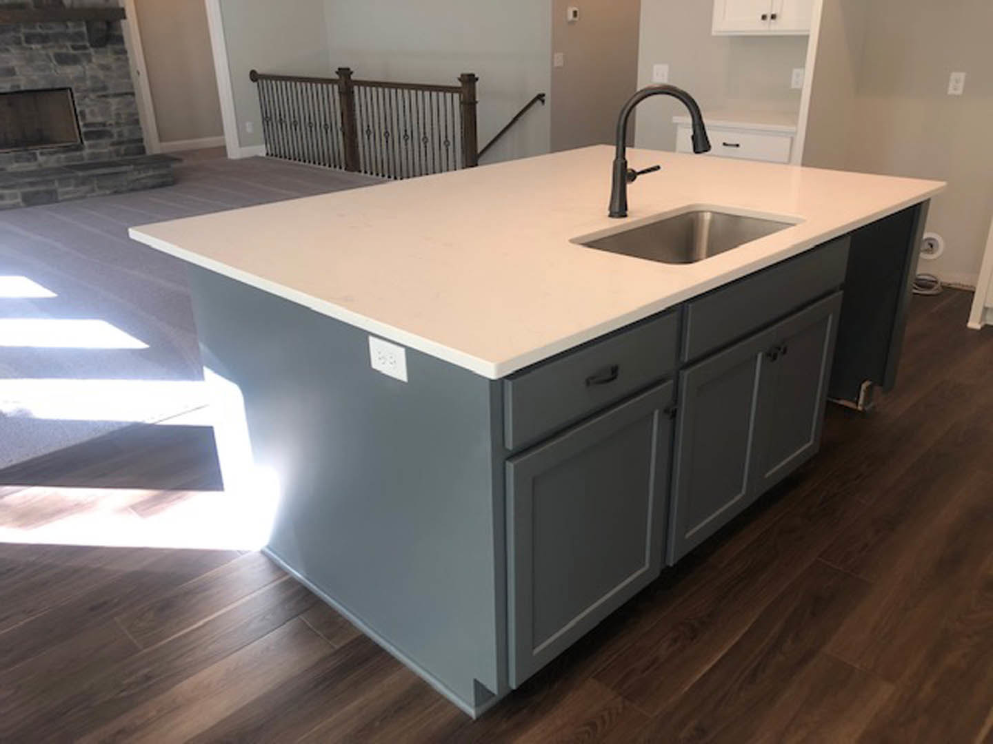White quartz kitchen island with integrated sink and matte black faucet, surrounded by light wood cabinetry and stone fireplace wall in the background