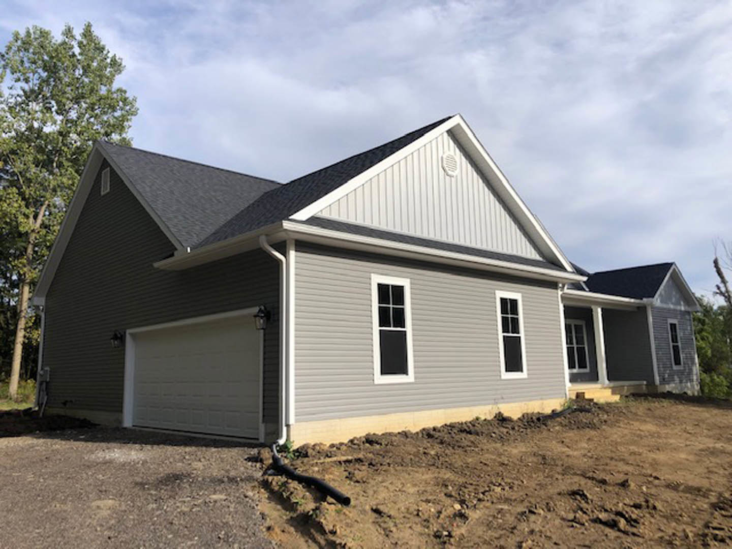 Two-story home with white-framed windows, light gray siding, attached garage with exterior light, dirt hill in foreground, cloudy sky above
