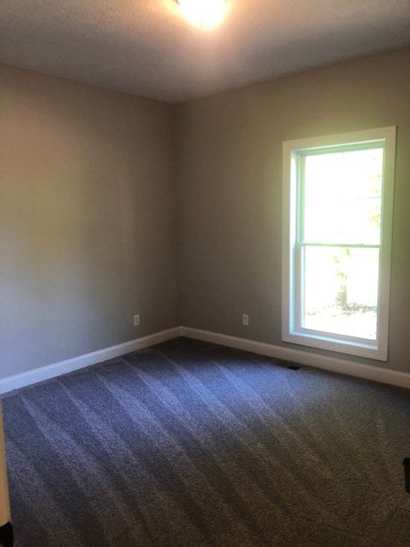 Carpeted bedroom with white-framed window, neutral walls, and ceiling light fixture