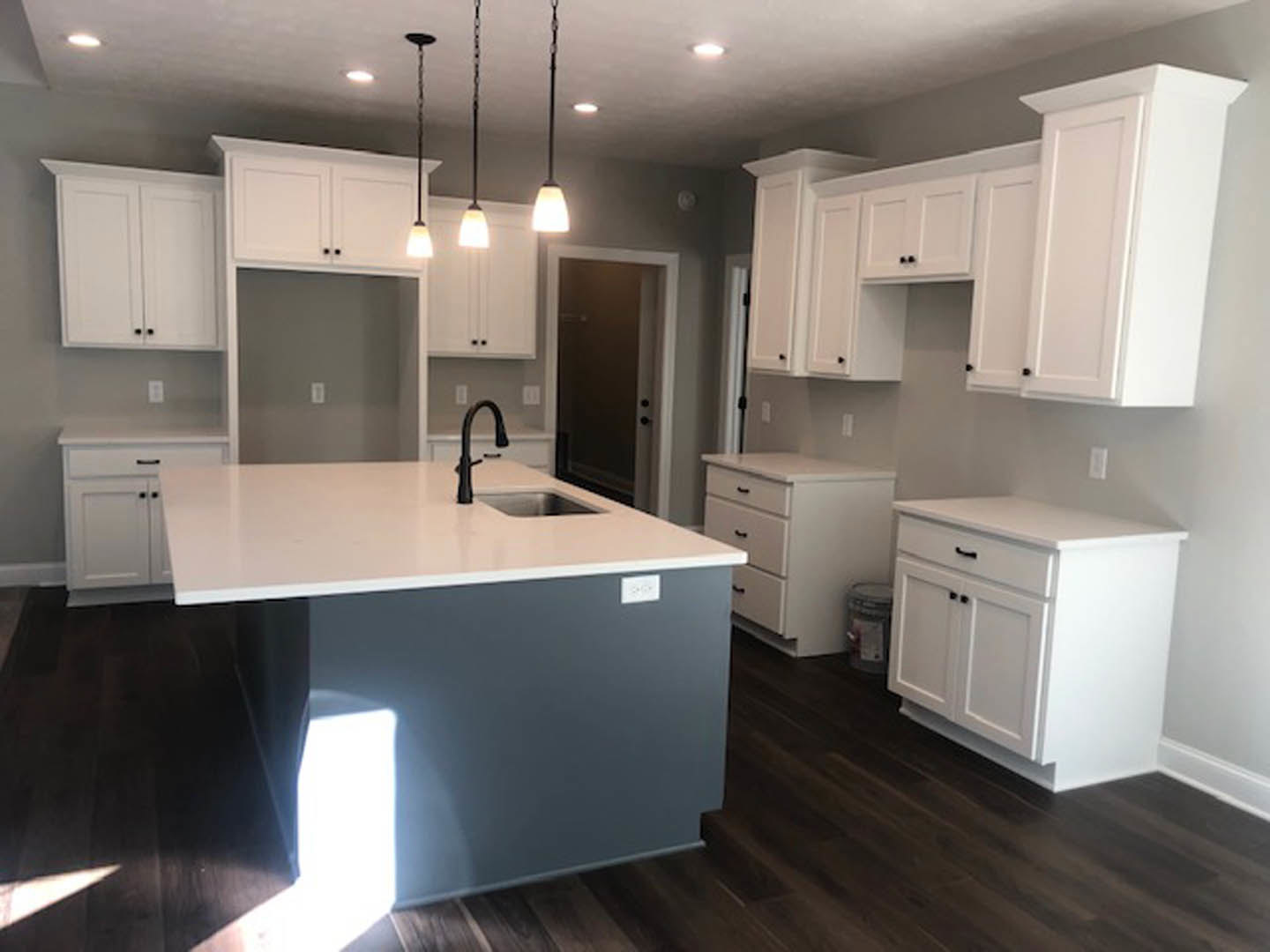 White kitchen with shaker cabinets, black handles, central island featuring a built-in sink and faucet, pendant light fixture above, hardwood flooring, and stainless steel