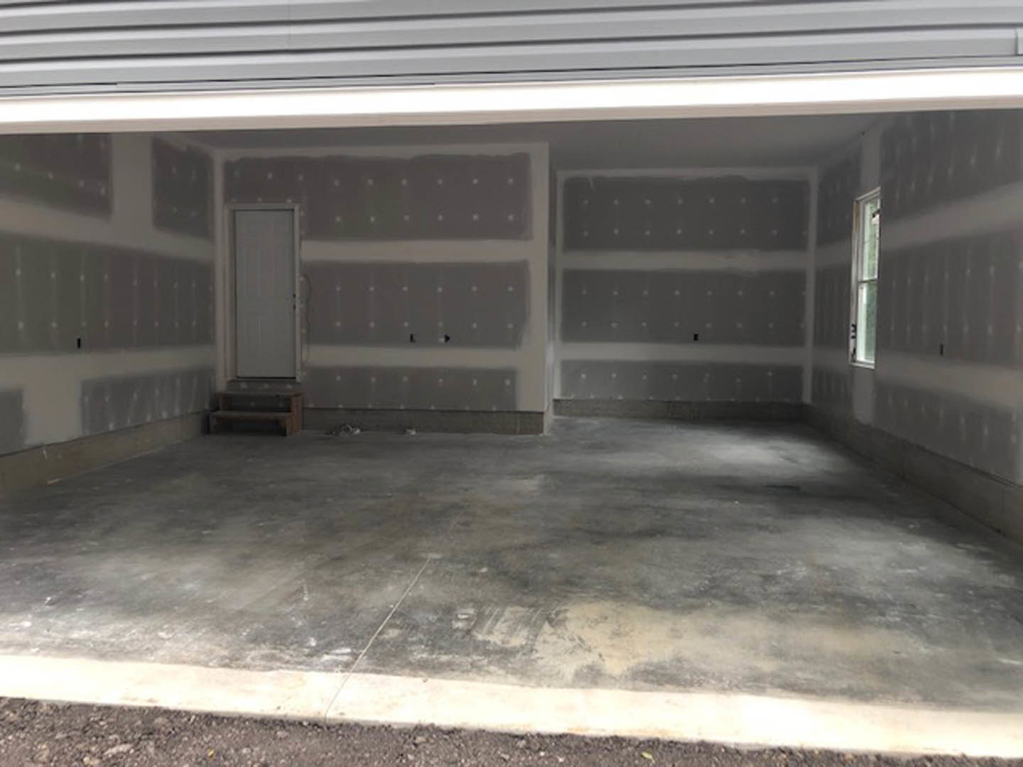 Garage interior with concrete floor, white door with black handle, window with white frame, wall showing holes, and shelf in background.