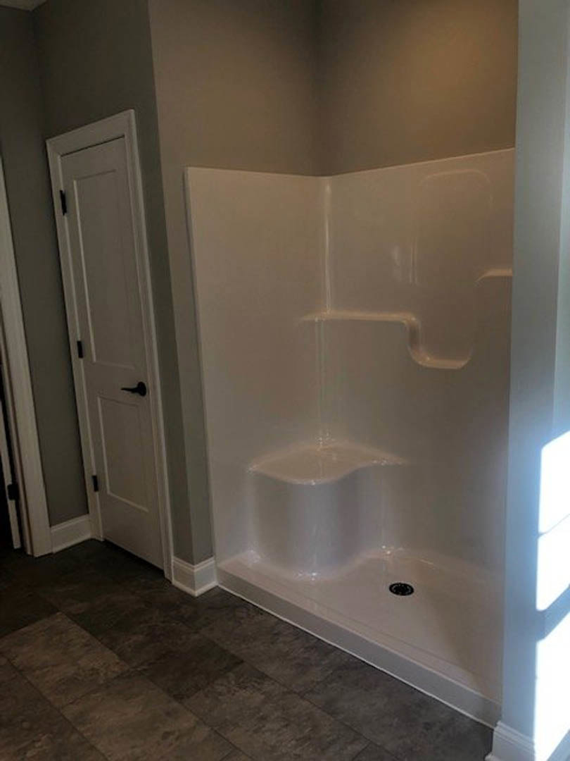 Walk-in shower with large gray floor tiles, linear drain, white walls, and a white door featuring a black handle