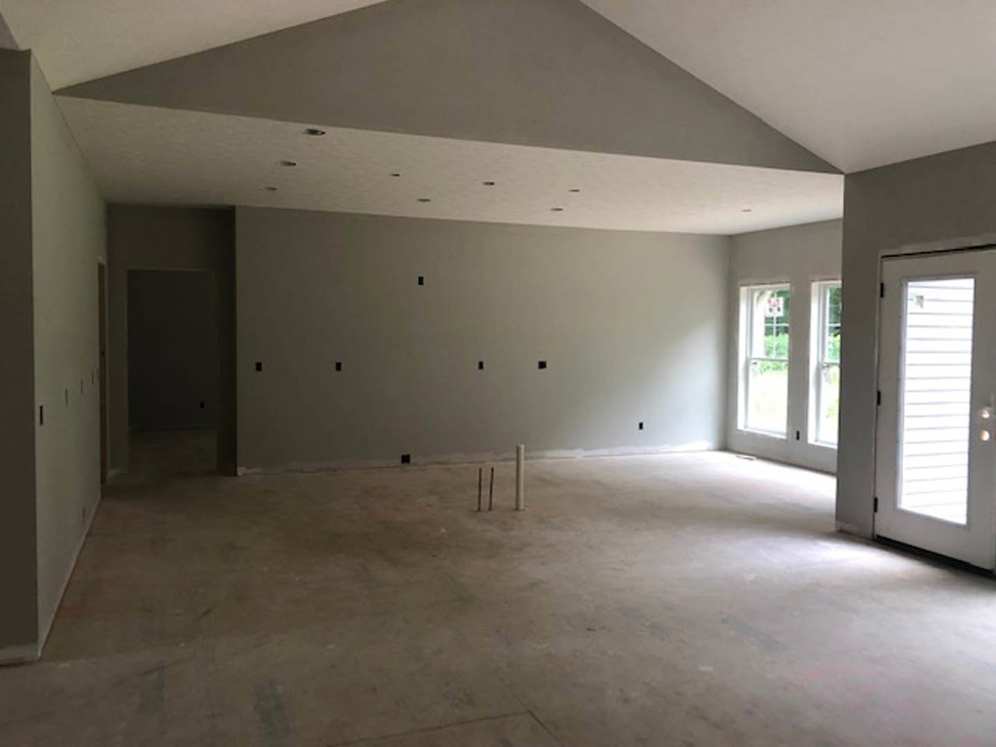 Concrete floor with exposed pipes, black accent wall featuring electrical outlets, white door with blinds, large windows framed by plaster walls, minimal indoor lighting.