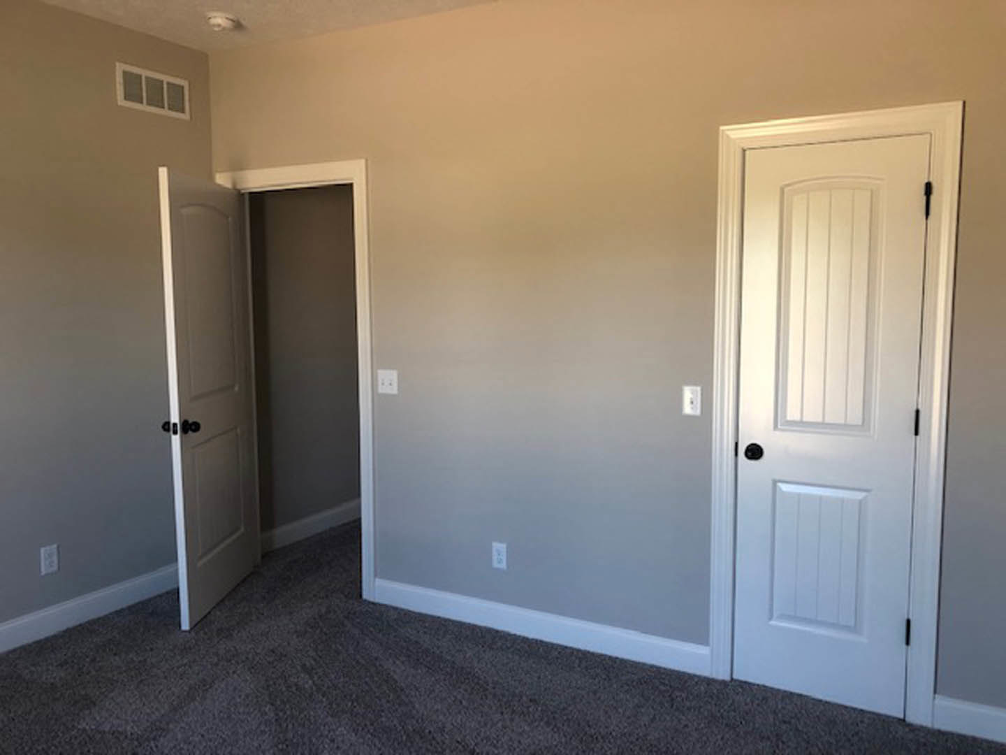 White walls and grey carpeted floor in a room featuring two doors with black handles, a window with a vent, and minimal interior finishes.