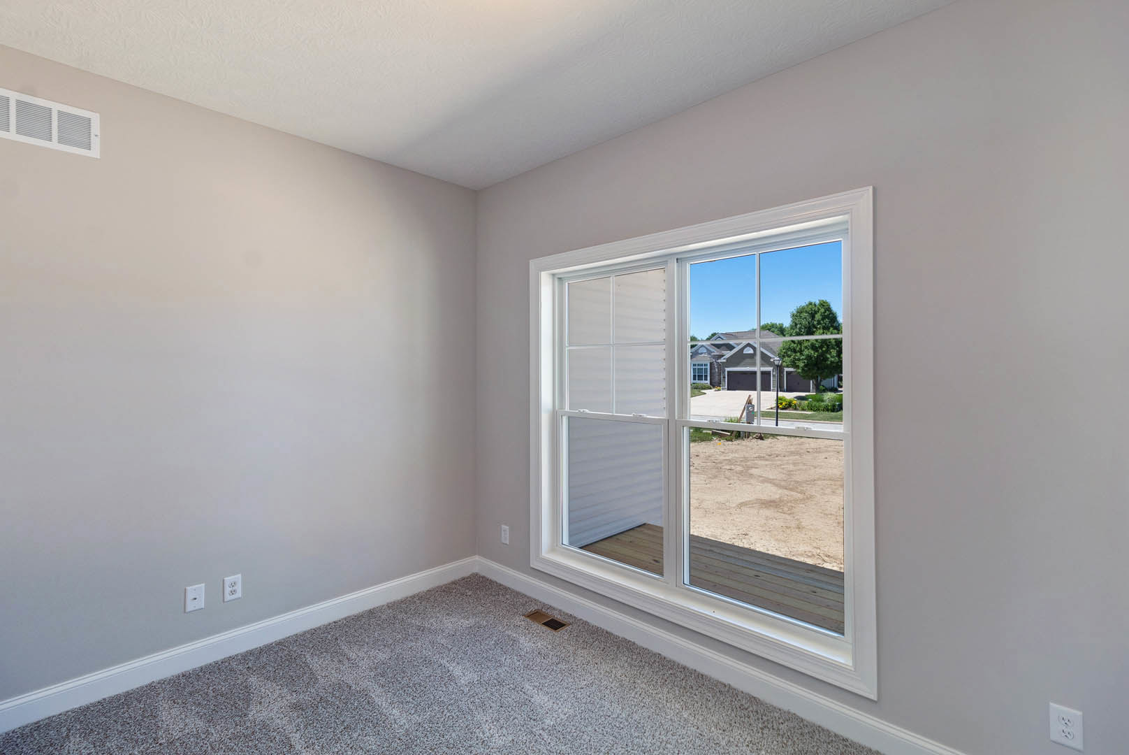 Carpeted room with white walls, large window overlooking a house and yard, wall outlet and air vent visible