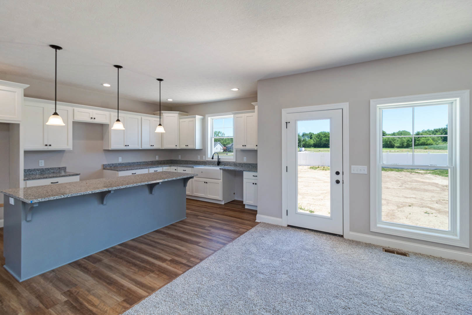 Open kitchen and living room featuring white cabinetry, wood flooring, light countertops, a window overlooking a dirt road and trees, and a door with a view of a field; black