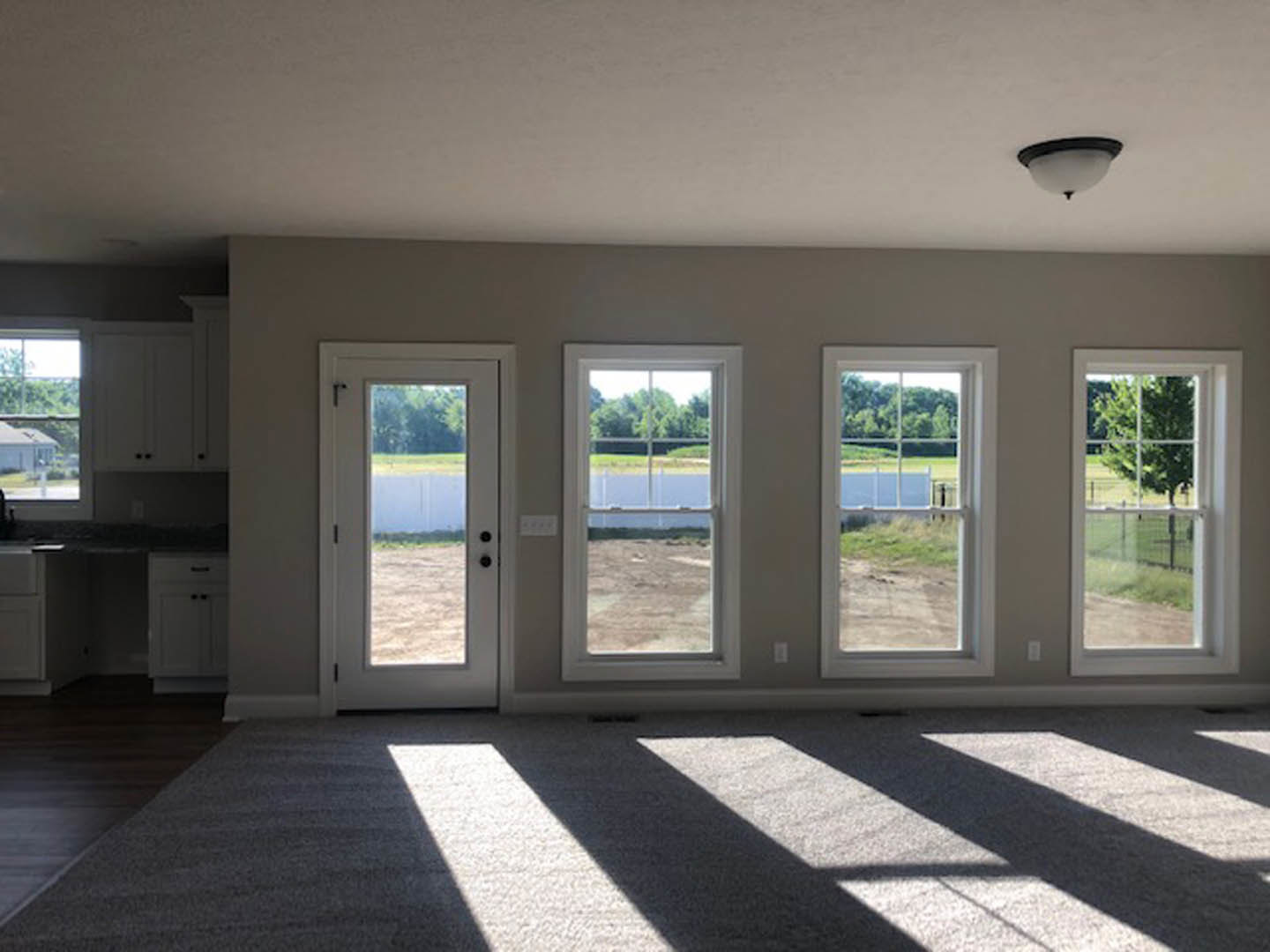 Bright room with large windows, glass door, light wood flooring, white walls, and views of grassy field, trees, and backyard fence
