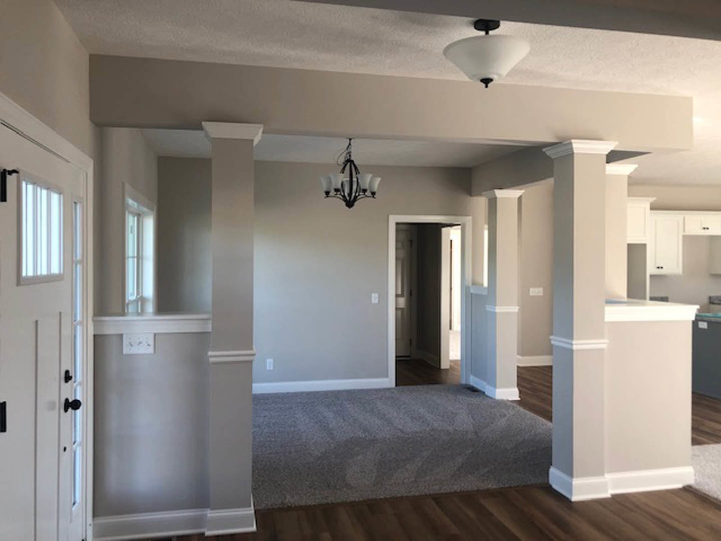 Wood flooring and white pillars in a spacious room, chandelier hanging from the ceiling, grey carpeted bench near a white door with window, open hallway leading to another area.