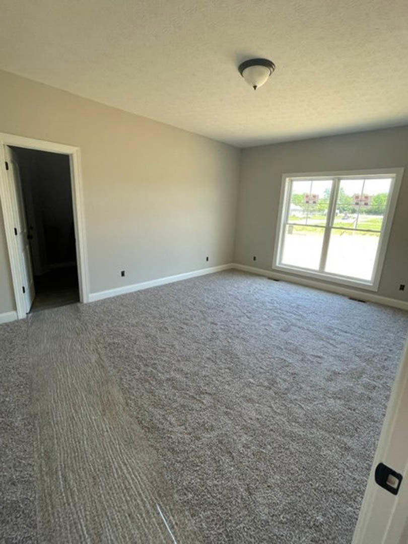 Carpeted bedroom with large window, white walls, ceiling light fixture, and doorway leading to a darker adjacent room