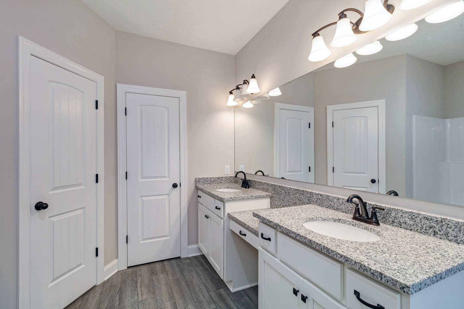 Bathroom with double white sinks set in a black and white speckled countertop, large wall mirror above, white cabinetry with black handles, and white door with black hardware.