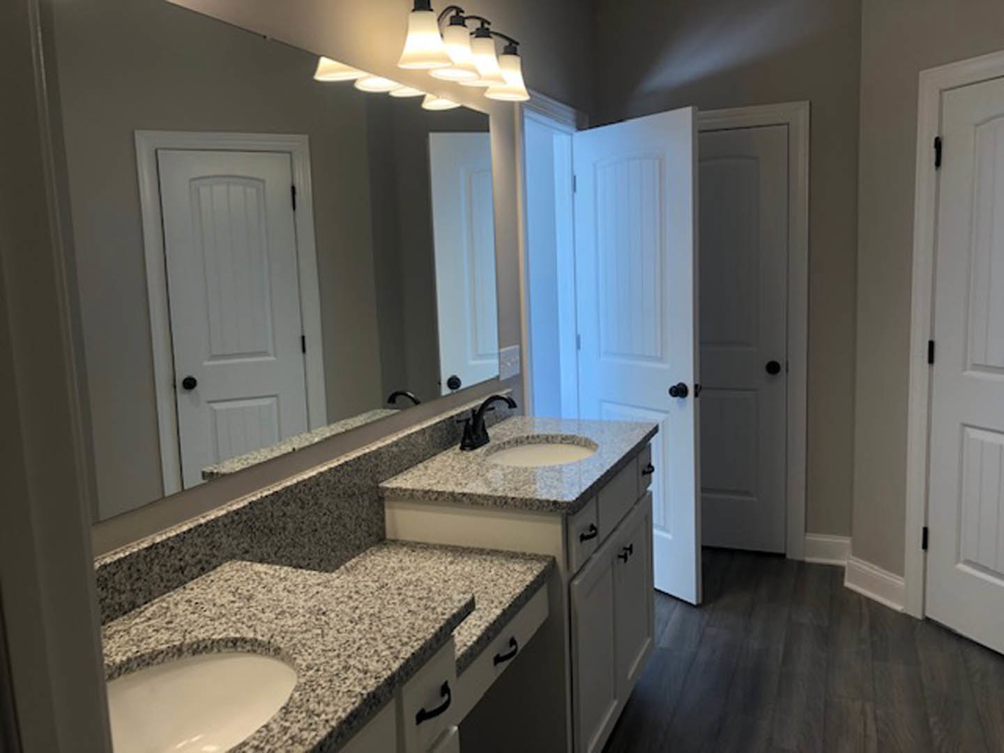 Bathroom with double vanity featuring white sinks, wide mirror above, dark wood flooring with white trim, white door with black hinge, and ceiling row of lights.