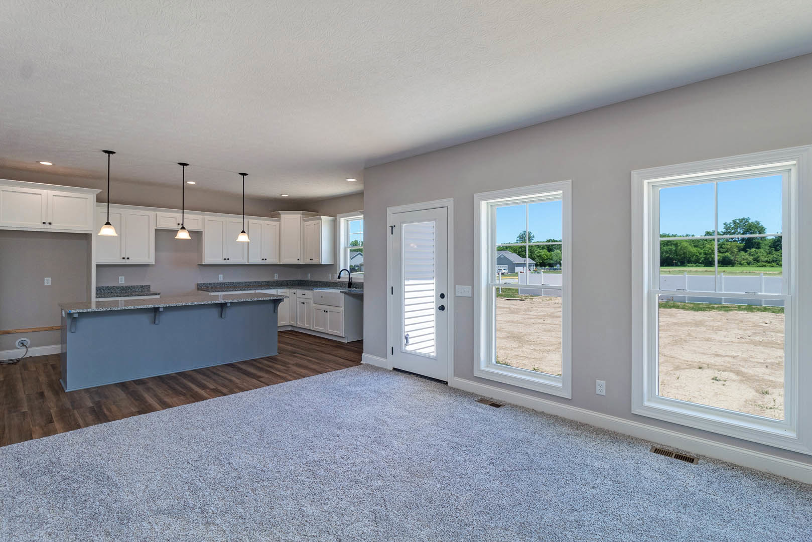 Open-concept kitchen with a large central island, wood flooring, white cabinetry, stainless steel sink, window overlooking a field and trees, white door with blinds, and adjacent