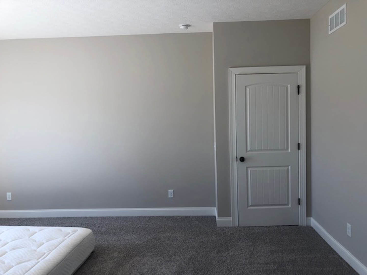 White paneled door with black knobs set in a white wall, carpeted flooring, mattress partially visible, window with vent above.