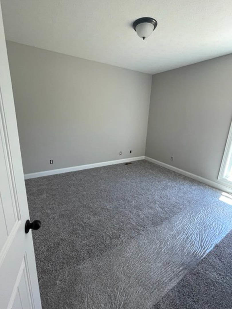 Neutral-toned carpeted room with white walls, ceiling fan, and white door featuring a silver knob; water pooled on floor near grey baseboard.