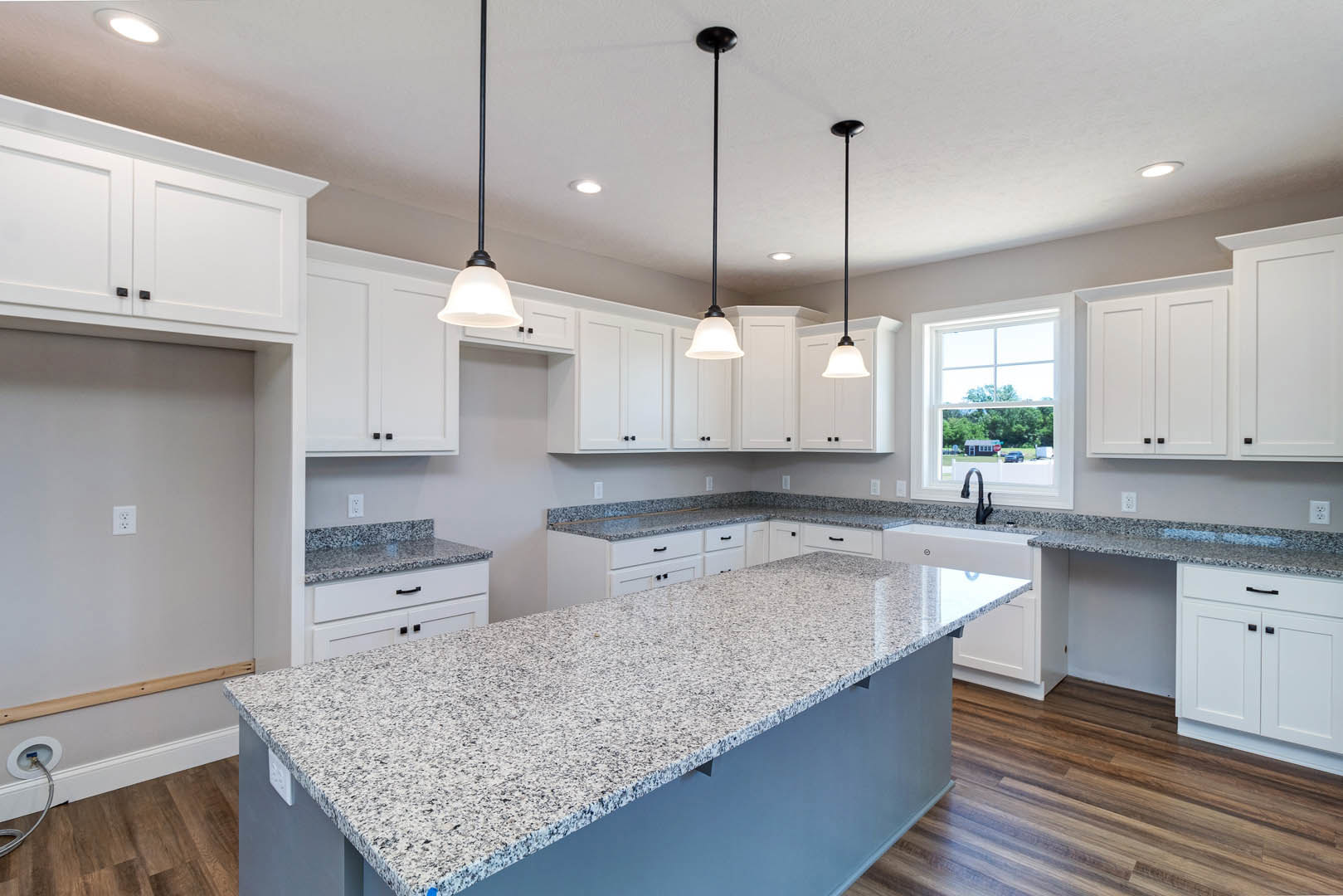White kitchen cabinets and black-and-white speckled granite countertops, stainless steel sink, modern appliances, light-colored walls, window overlooking road and trees.