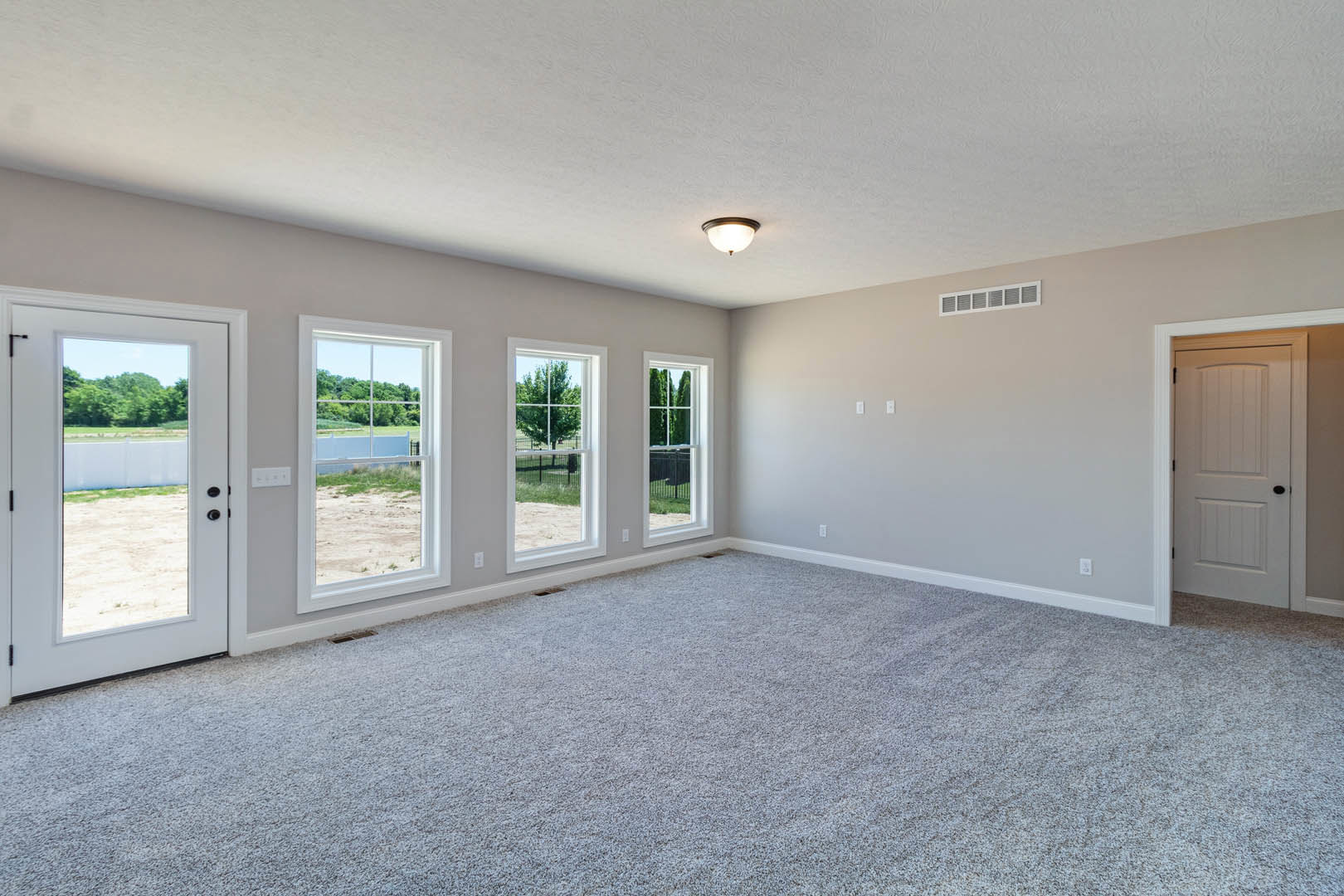 Carpeted room with large windows overlooking a field and trees, white door with black knob and glass panel, ceiling light fixture, pale walls