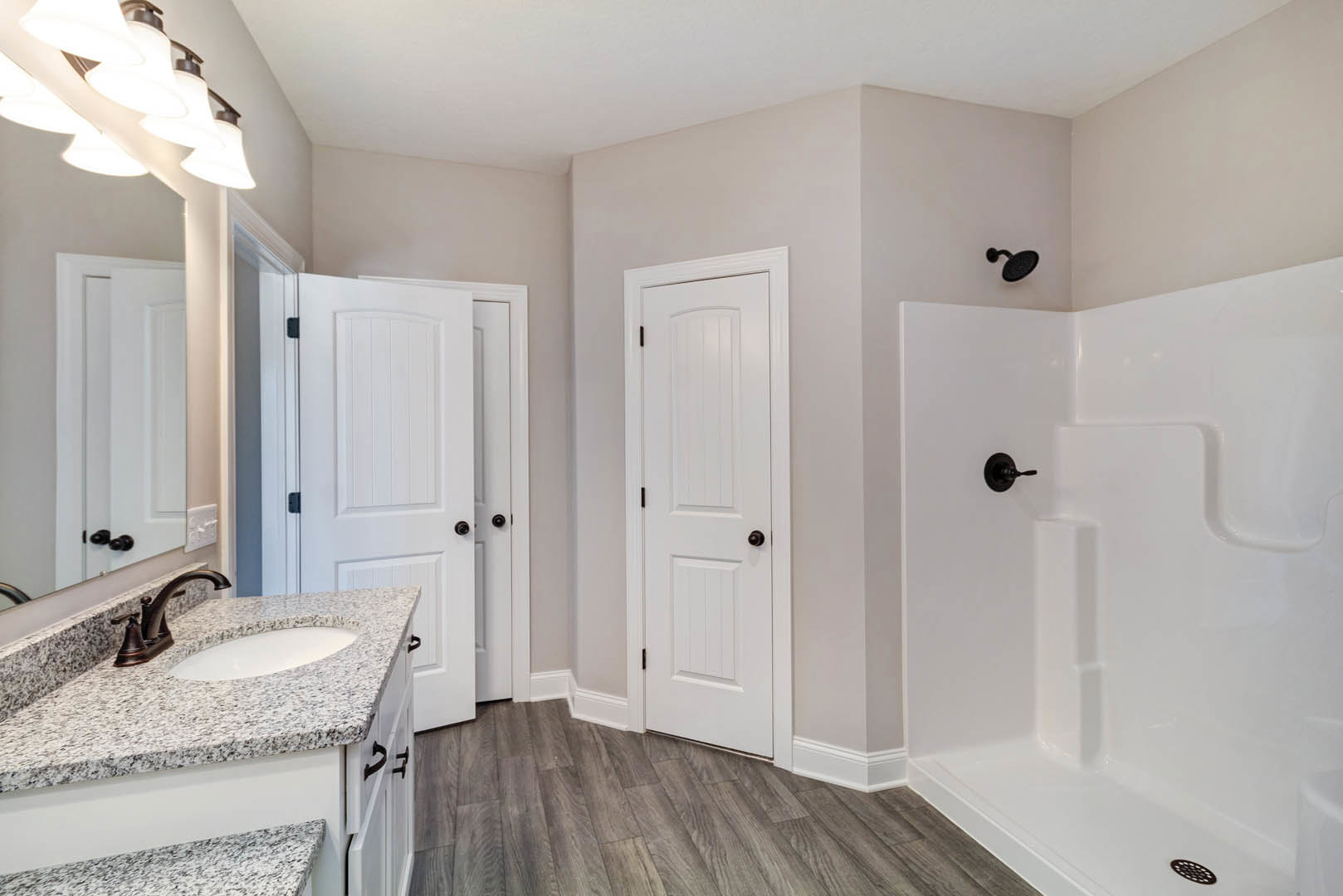 Bathroom with white door and black knob, white shower featuring black handle, light fixture above sink and countertop, neutral cabinetry, and tile flooring.