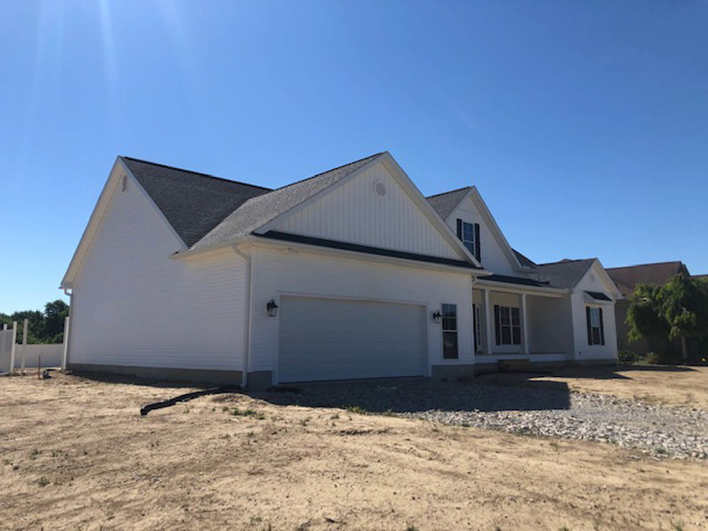White two-story house with attached garage, gray roof, and large windows, surrounded by trees and dirt lot under clear sky