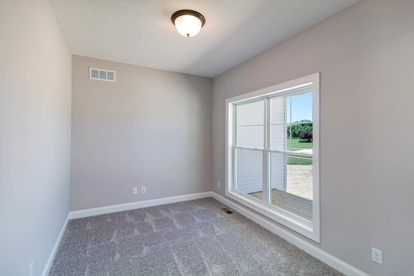 Grey carpeted floor, white walls with base molding, ceiling light fixture, wall outlet and vent, large window overlooking green lawn