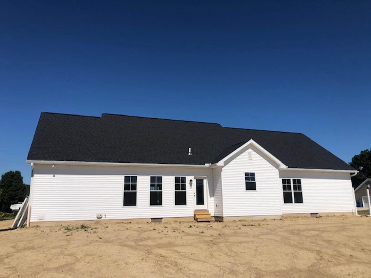 White siding house with black shingle roof, square-paned windows with white frames, surrounded by dirt field and trees under blue sky