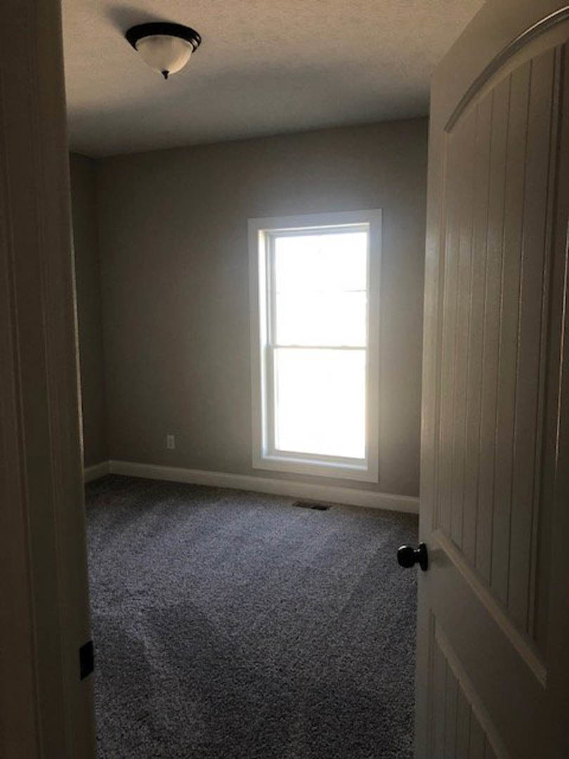 Bedroom with beige carpet, white walls, large window letting in natural light, black door knob, and ceiling light fixture