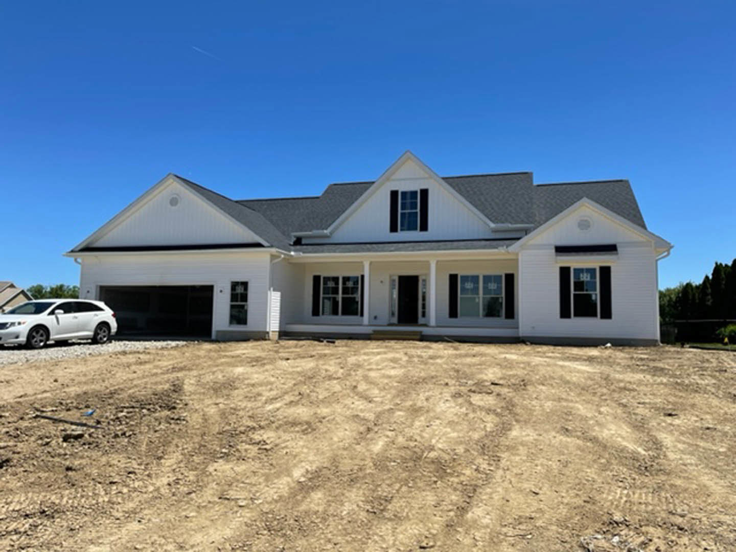 White two-story house with white-framed windows, covered front porch, and a white car parked on a dirt driveway under a clear blue sky.