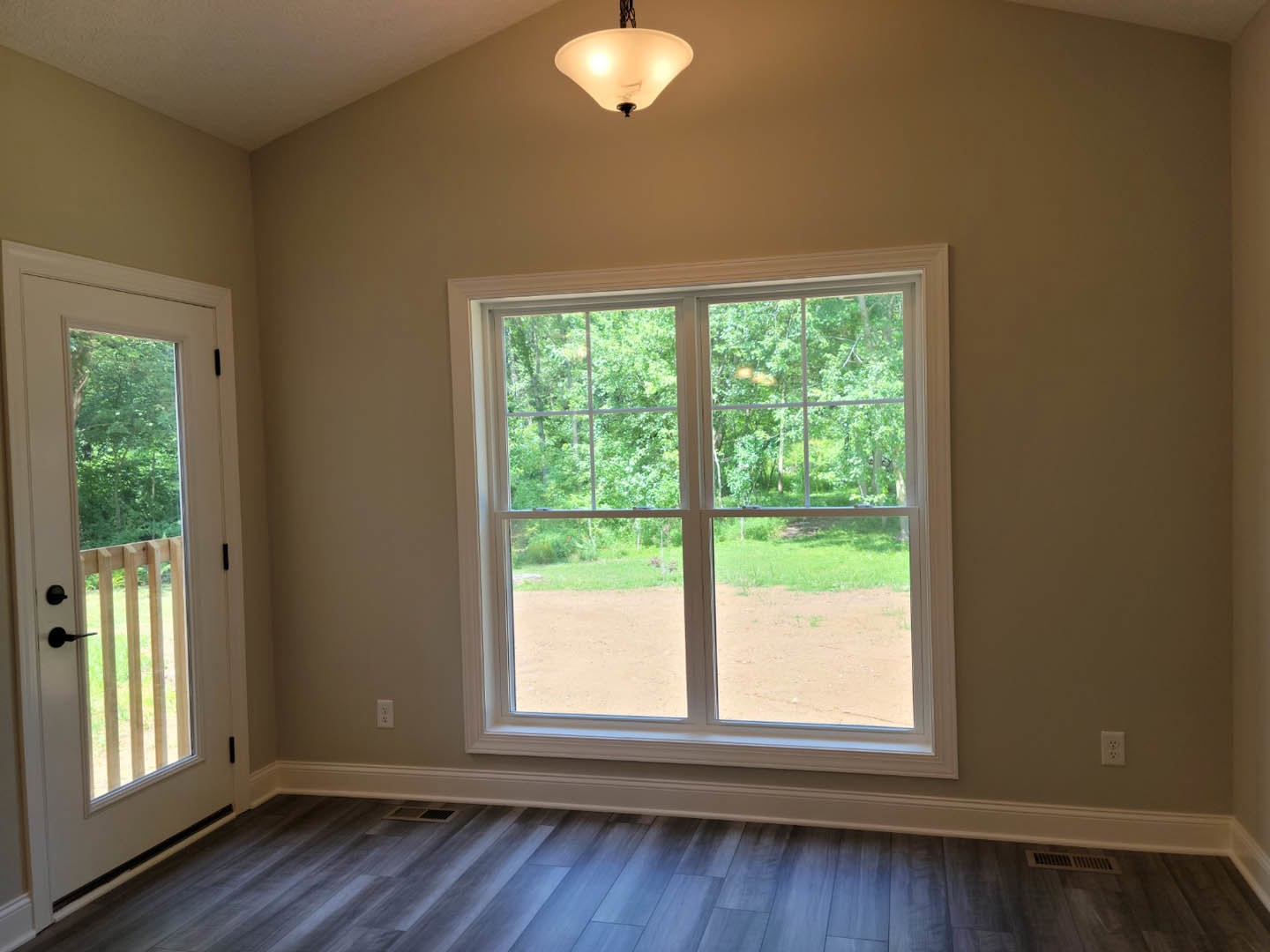 Sunlit room featuring a large window overlooking trees, wood flooring with white baseboards, glass door leading outside to a fenced area, and ceiling light fixture with a white
