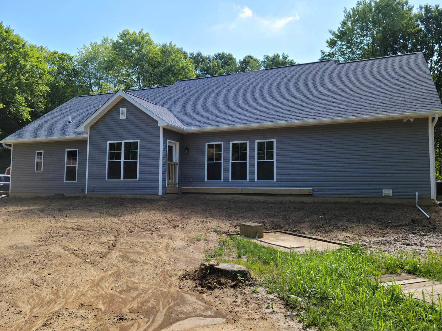 Two-story house with white-framed windows and covered porch, surrounded by dirt and patches of grass, trees and blue sky with clouds in the background, concrete block in