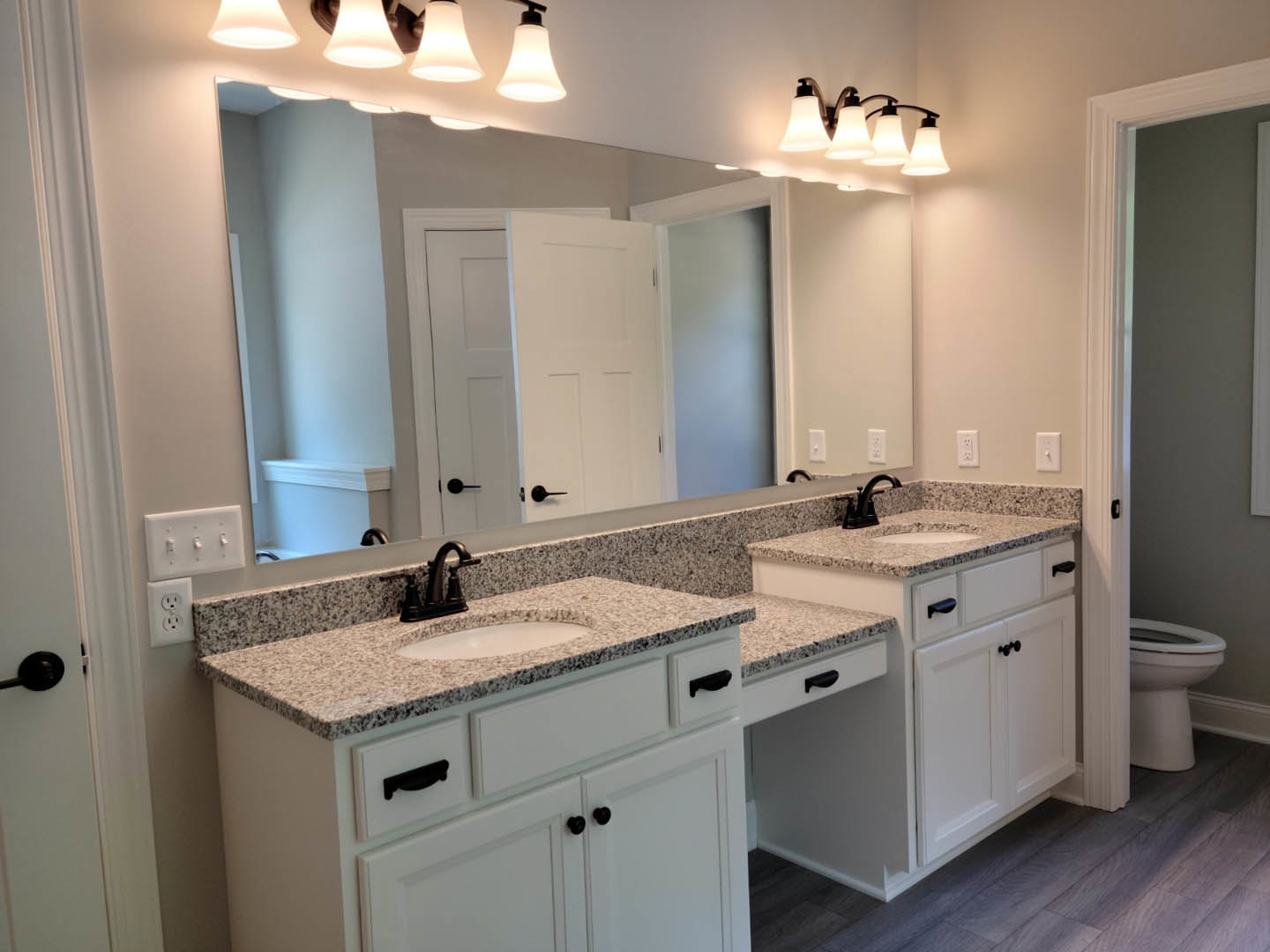 Bathroom with expansive wall mirror, double white sinks set in a stone countertop, modern chrome faucets, light tile flooring, and dark cabinetry beneath sinks.
