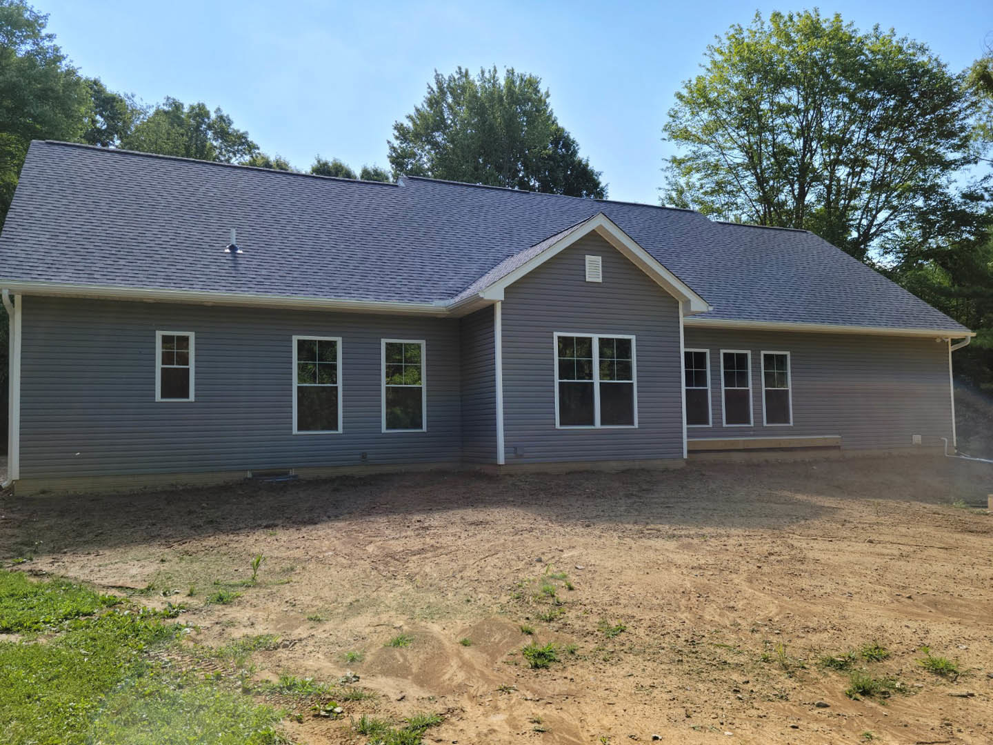 Two-story house with white-trimmed windows, gray siding, and gabled roof, surrounded by dirt field and scattered trees; small white cottage visible in the background.