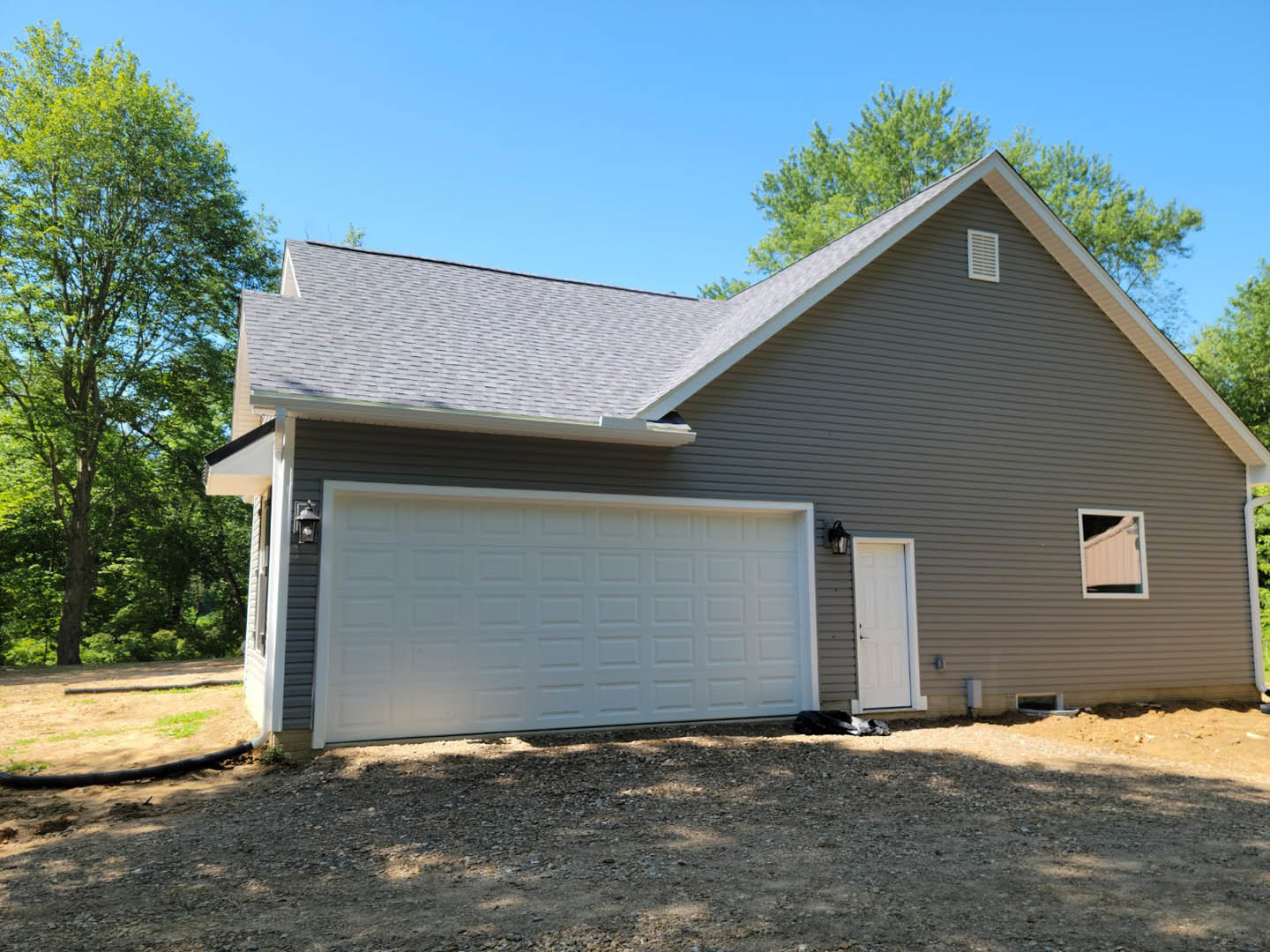 Two-story home with gray siding, attached single-car garage featuring a white paneled door, large front window, and leafy tree partially shading the driveway