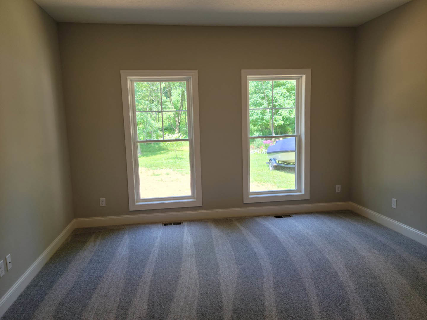 Carpeted room with two windows, white walls, floor vent, and a view of trees and a boat on a trailer outside