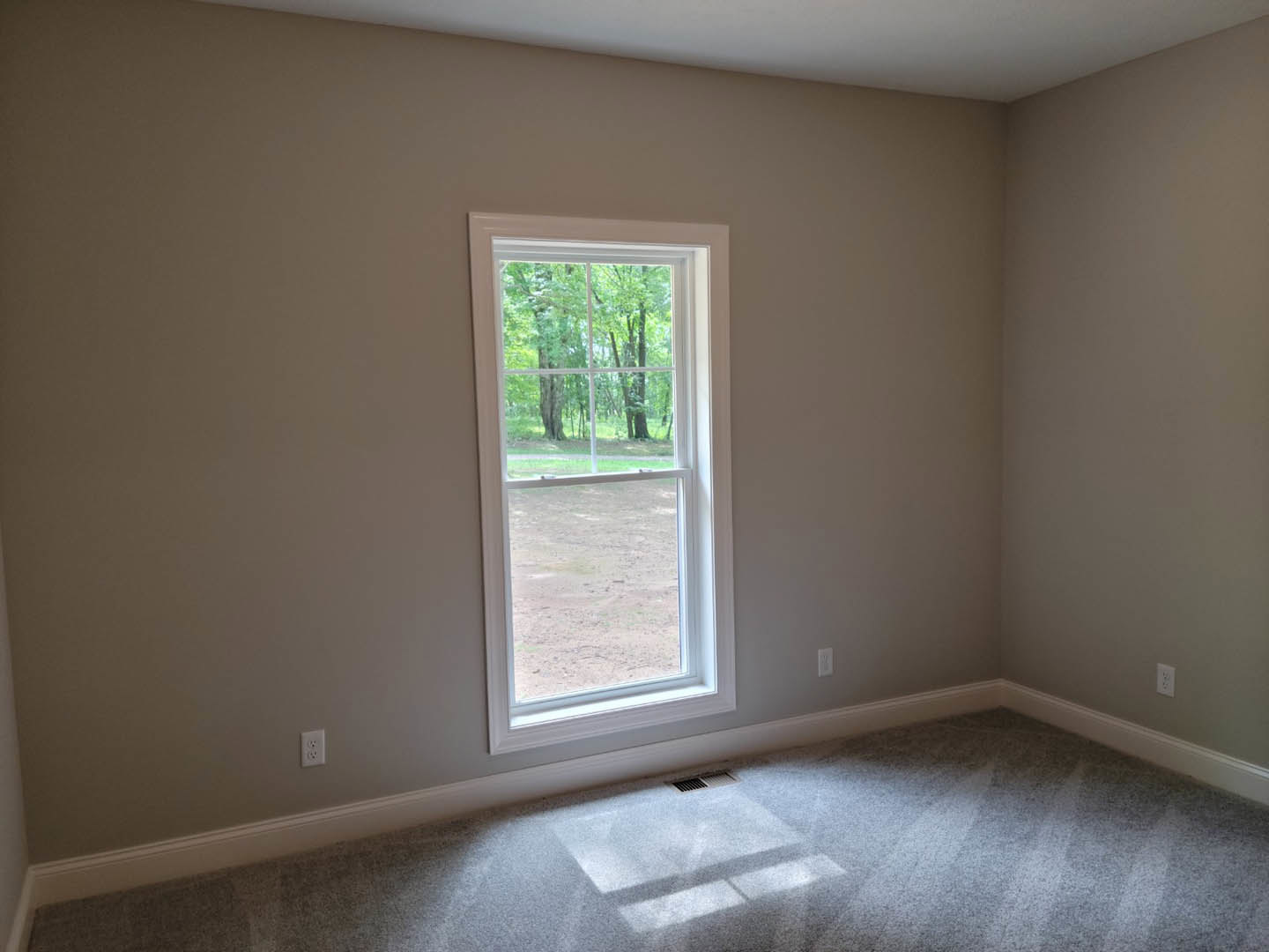 White carpeted floor with rectangular metal vent, large window framed in white trim, plaster walls, leafy trees visible outside