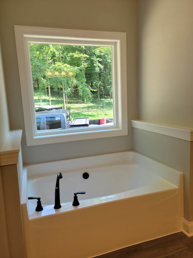 Freestanding white bathtub with chrome faucet and black handle beneath large window, white walls with molding, leafy trees visible outside