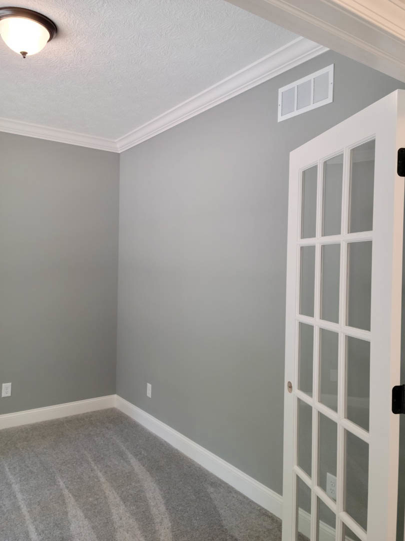 Carpeted room with white baseboards, white paneled door, ceiling light fixture, wall vent, and window