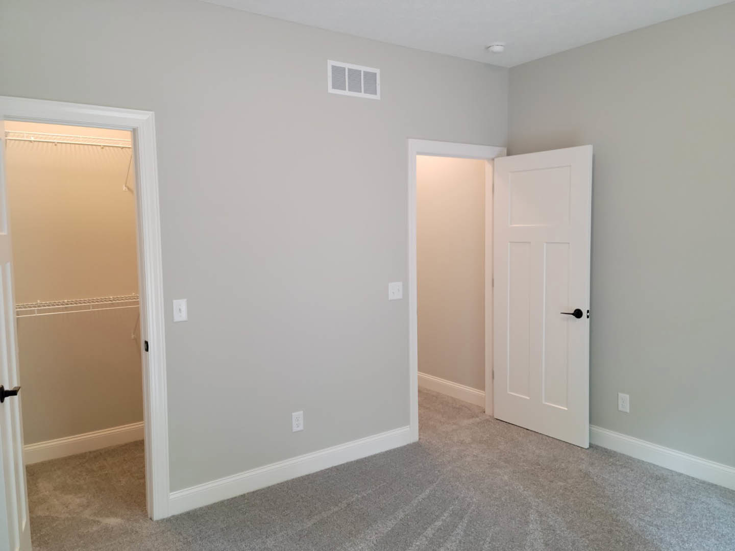 Bedroom with open white door featuring black handle, white closet with built-in shelf, carpeted floor, white walls, electrical outlets, and wall vent.