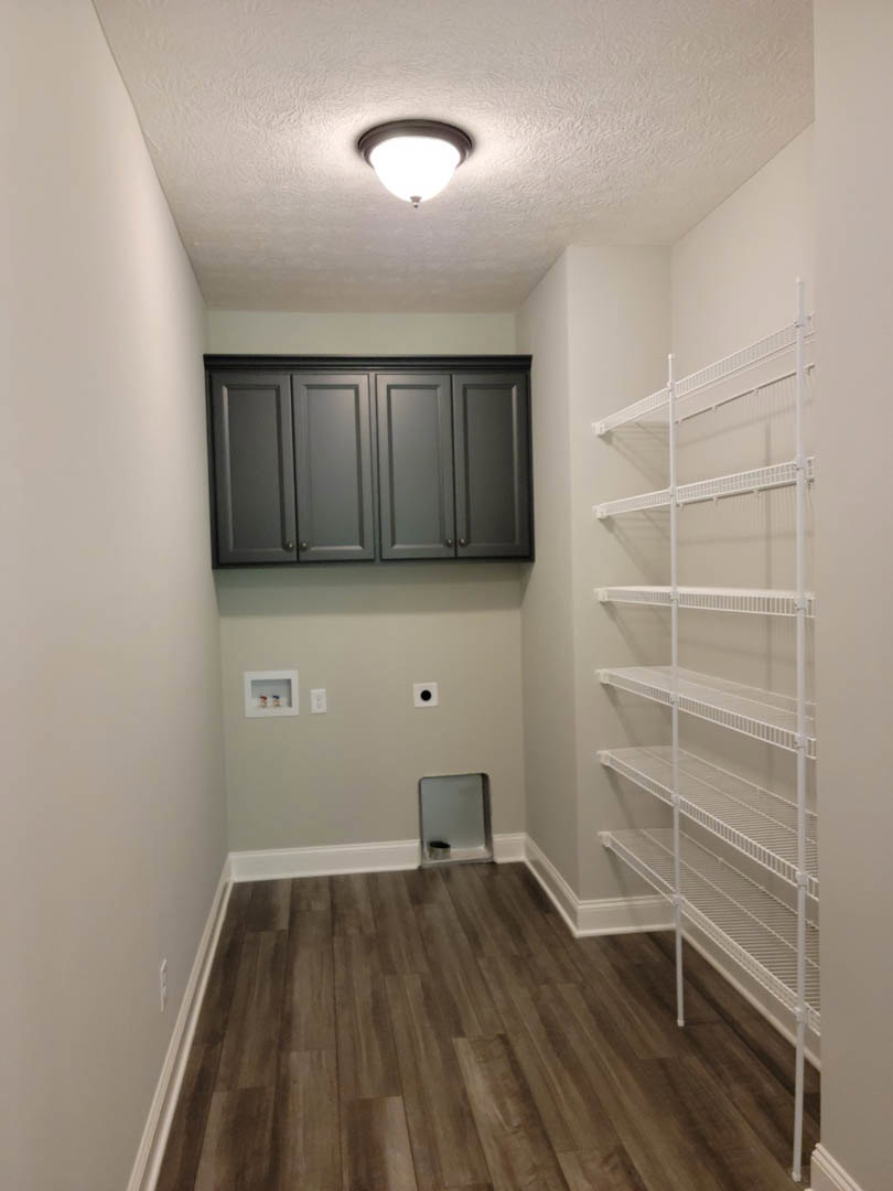 Wood flooring with white trim, built-in white shelves along the wall, grey cabinetry, plaster ceiling with a modern light fixture, and a white rectangular storage container with a