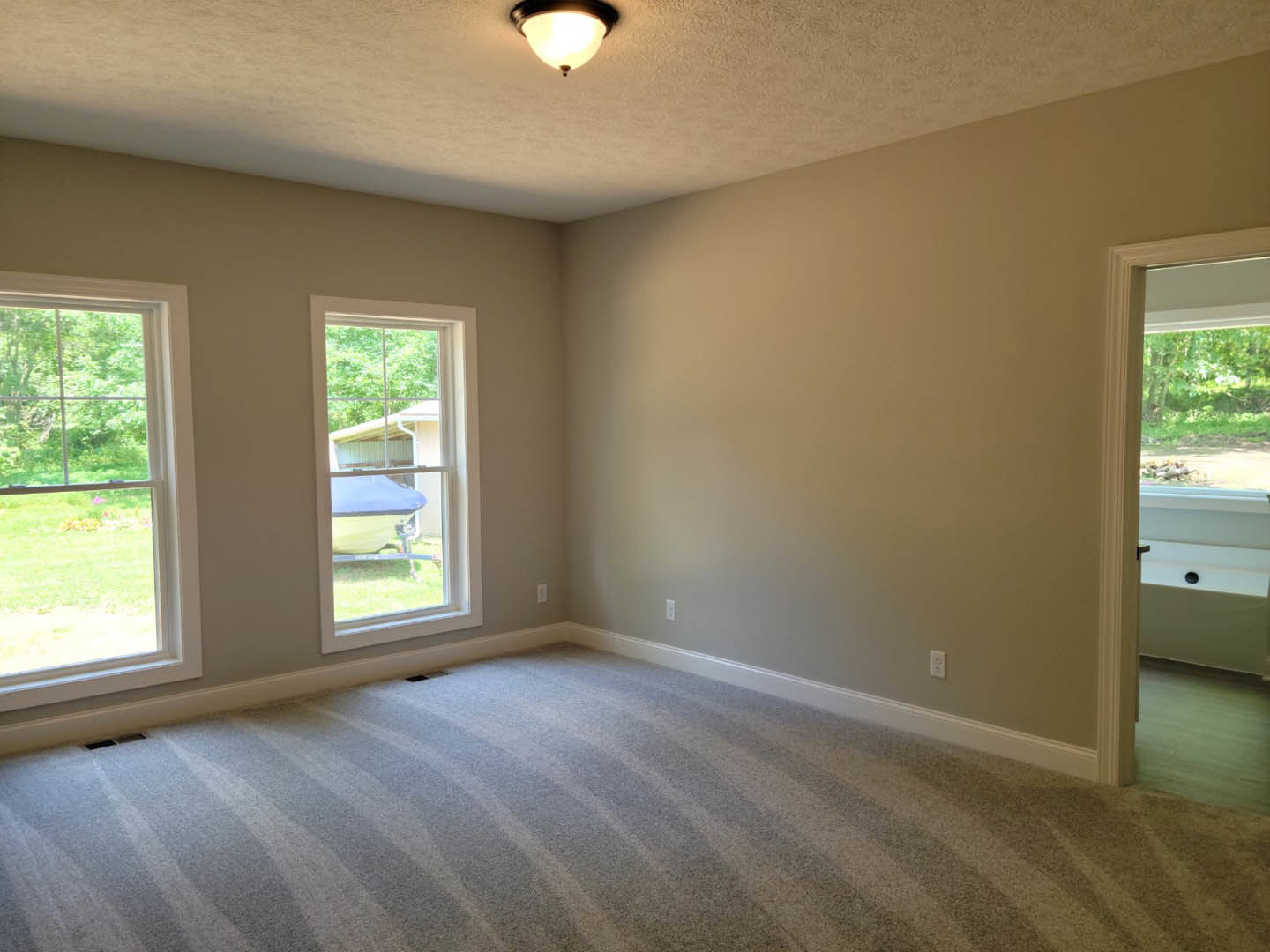 Carpeted room with white walls, two windows showing a grassy field and a distant boat, ceiling light fixture, and a door.