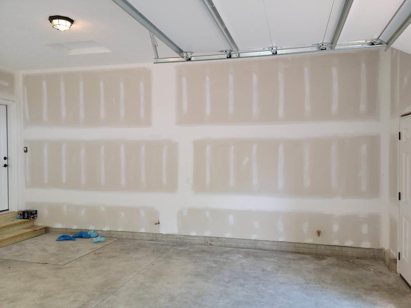 Concrete-floored room featuring a white-painted garage door, plaster walls, ceiling light fixture, blue bag on the ground, and metal corner trim.