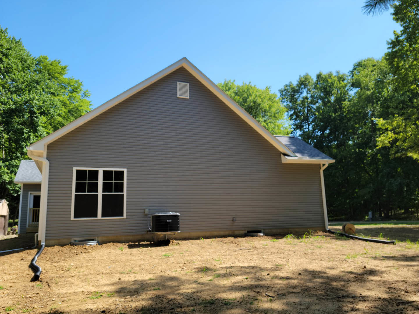 Two-story home with gray siding, multiple white-trimmed windows, gable roof, grassy yard bordered by dirt, mature trees in the background