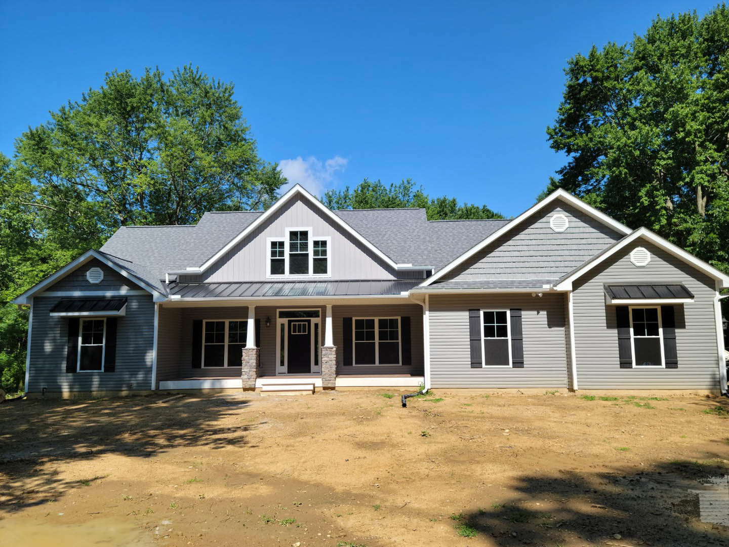 Two-story house with light siding, black front door with white trim, white-framed windows, covered porch, dirt yard, and mature trees in the background