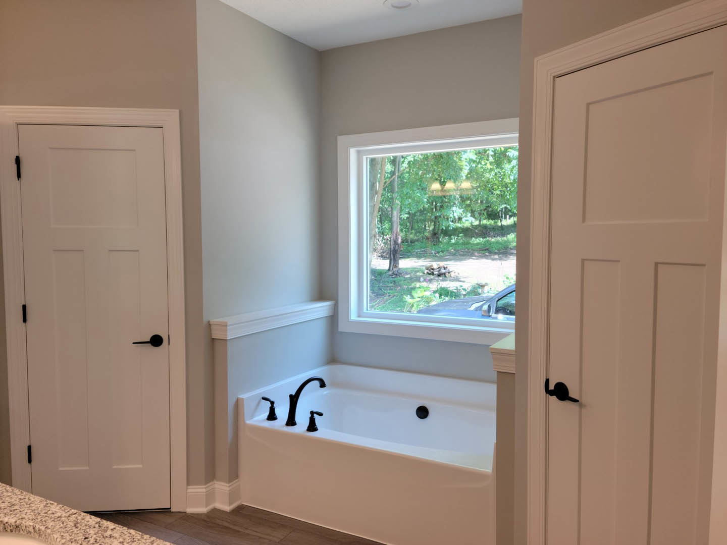 Freestanding white bathtub with chrome faucet and black handle, positioned near a window overlooking the outdoors, adjacent to a white door with black hardware in a modern