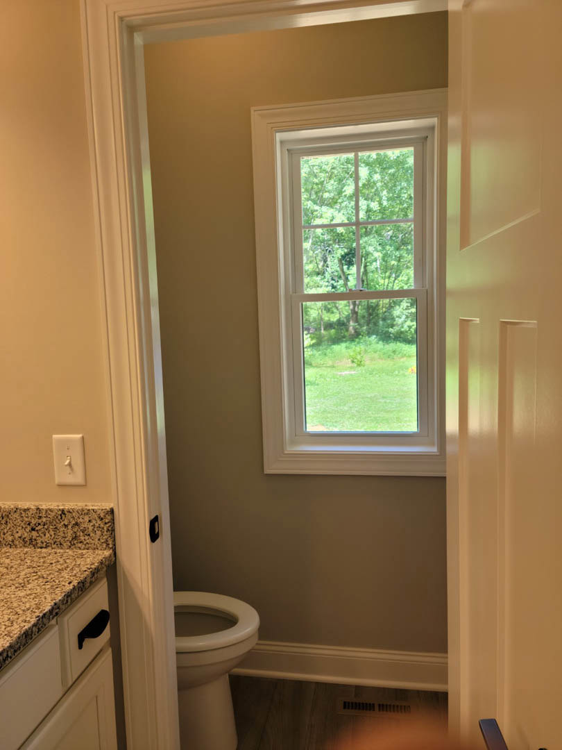 Bathroom with white tile walls, rectangular window overlooking green lawn, modern sink with chrome faucet, quartz countertop, toilet partially visible, and brushed metal light