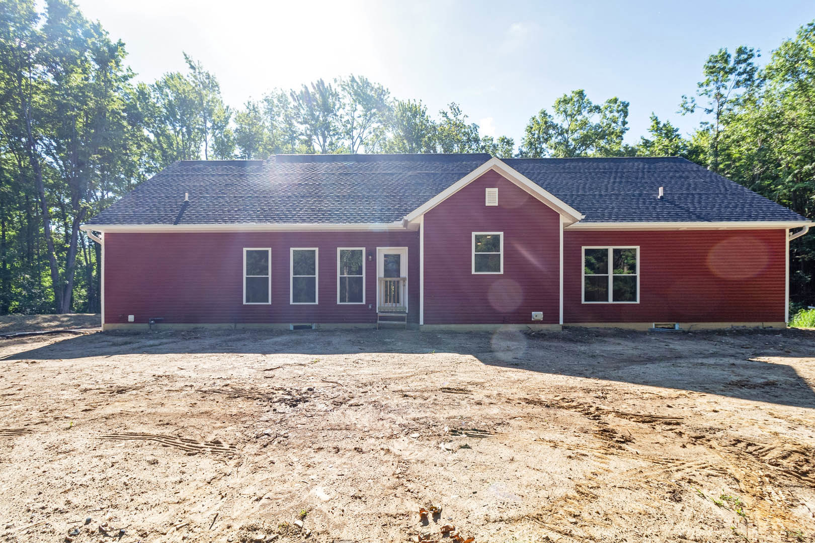 Partially built house with red exterior wall, white-framed window, dirt ground scattered with sticks, trees in background