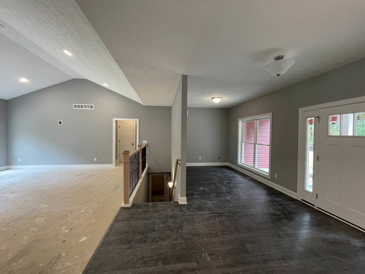 Dark wood flooring in a room with a white door featuring glass panes, a staircase with a close-up of the railing, pink blinds on a window, and a ceiling light fixture.