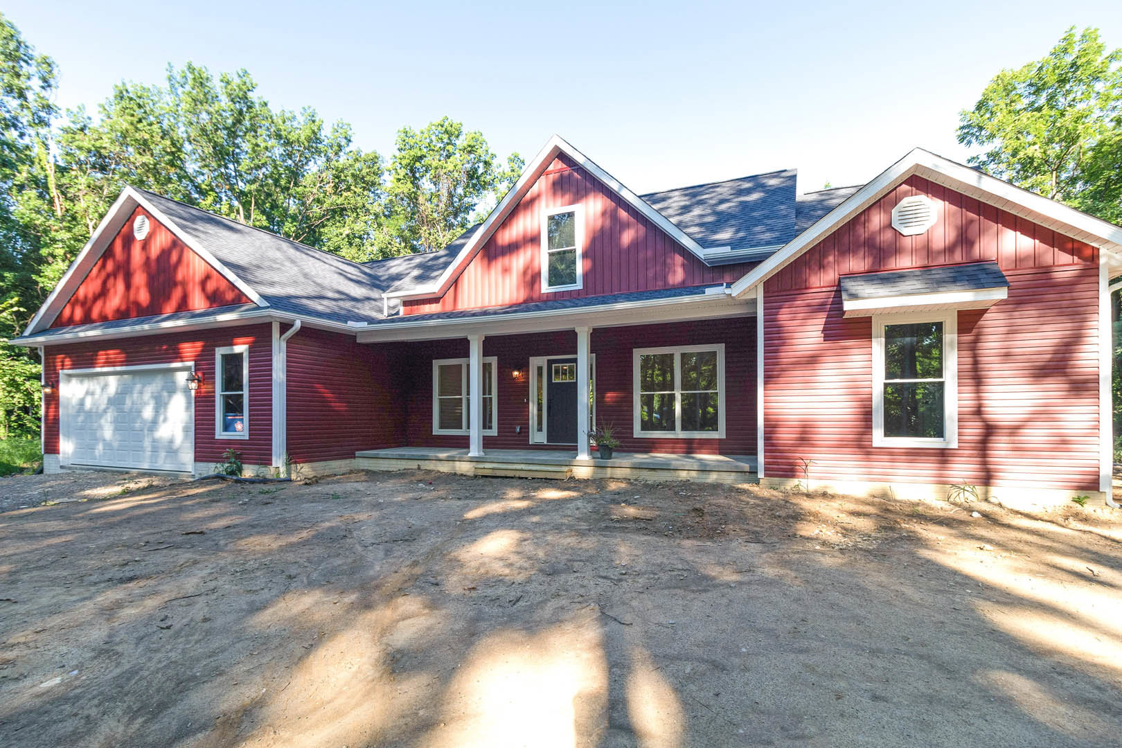 Red siding house with white trim, white garage door, red roof, white vent, window with white frame, red truck parked on dirt driveway, surrounded by trees under blue sky.